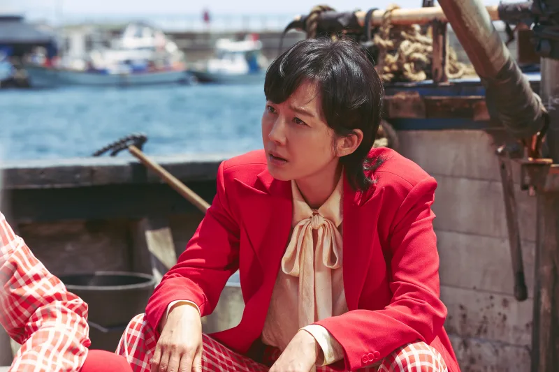 바다를 배경으로 배위에 빨강 정장을 입고 앉아 있는 영화 밀수의 배우 염정아